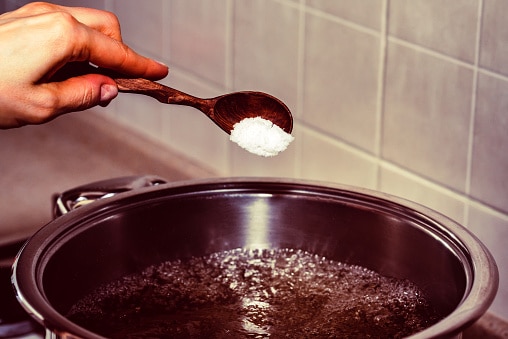 Close up hand with wooden spoon adds salt to boiling water in saucepan, preparing soup